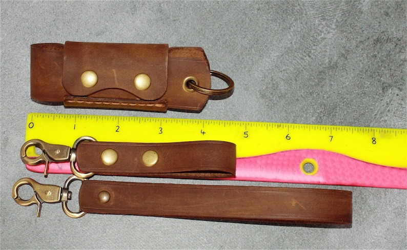 May include: Brown leather keychains and a key holder with brass hardware. The keychains have a swivel clasp and are secured with brass rivets. The key holder has a snap closure. A yellow and pink ruler is in the background.
