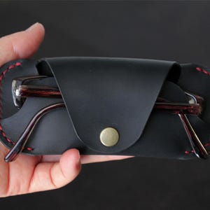 Puede incluir: Un estuche de cuero negro para gafas con una solapa plegable asegurada por un botón de presión de metal color latón. El estuche presenta costuras rojas visibles a lo largo de los bordes y contiene un par de gafas oscuras con patrón de madera.