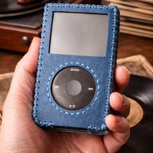 May include: A portable digital music player encased in a blue leather cover with visible cross-stitching along the edges. The device features a rectangular screen above a circular black click wheel with white text including MENU, play, pause, and skip icons.