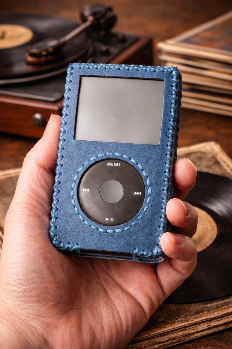 May include: A portable digital music player encased in a blue leather cover with visible cross-stitching along the edges. The device features a rectangular screen above a circular black click wheel with white text including MENU, play, pause, and skip icons.