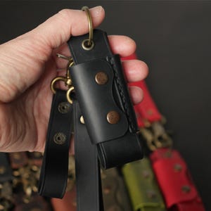 May include: Black leather keychain with a brass ring and clasp. The keychain features a small, riveted leather sheath. Additional keychains in red, green, and other colors are visible in the background.