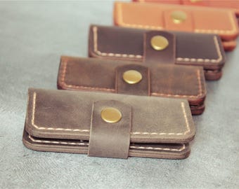 May include: A collection of leather wallets in various shades of brown, from light tan to dark brown. Each wallet features a flap secured with a gold-colored button. The wallets are stacked and have white stitching.