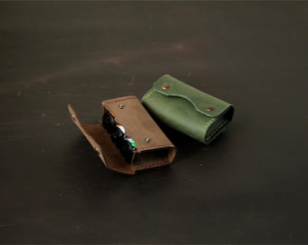 May include: Two rectangular leather cases with snap closures. One case is dark brown and open, revealing internal contents, while the other is forest green and closed. Both feature visible stitching along the edges and metallic hardware fasteners.