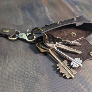 May include: A black leather key holder with a strap and metal hardware. Several keys are attached to the holder. The key holder is open, revealing the interior. The background is a dark, textured surface.