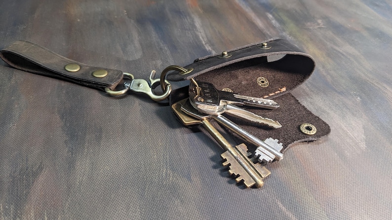 May include: A black leather key holder with a strap and metal hardware. Several keys are attached to the holder. The key holder is open, revealing the interior. The background is a dark, textured surface.