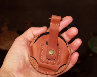 May include: A circular tan leather holder featuring a vertical strap with a brass-colored metal snap button closure. The leather case has visible stitching along the edges and a small loop at the top, designed to secure a small round object inside.