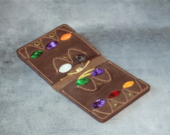 May include: A brown leather wallet with decorative stitching and colorful gemstone accents. The wallet is open, revealing multiple compartments and a variety of gemstones in shades of green, purple, orange, and red. The wallet is secured with brass snap closures.