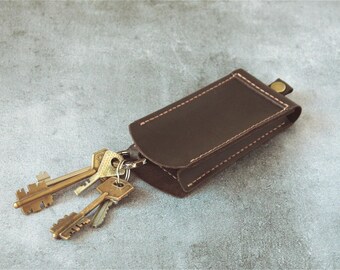 May include: A brown leather key holder with a rectangular shape and a curved flap. The key holder is stitched with white thread and has a small brass button closure. Several brass keys are attached to a metal ring.