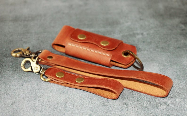 May include: A set of three brown leather accessories: a key fob, a wrist strap, and a small case. The key fob and wrist strap have brass-colored clasps and rivets. The case has a snap closure and a key ring.