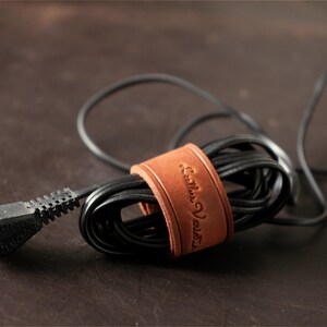 May include: A tan leather cable organizer strap wrapped around a coiled black electrical cord. The leather strap features the embossed text Leather Varsity and is secured with a metal snap button, keeping the black cable neatly bundled together.
