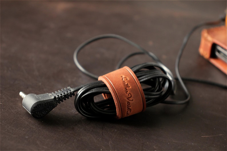 May include: A tan leather cable organizer strap wrapped around a coiled black electrical cord. The leather strap features the embossed text Leather Varsity and is secured with a metal snap button, keeping the black cable neatly bundled together.