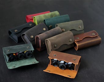 May include: A collection of small, rectangular leather cases with metal snap closures, arranged in various colors including red, green, brown, and black. Two open cases in the foreground reveal three cylindrical film canisters inside each.