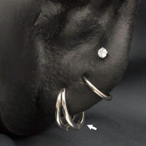 May include: A black ear with multiple piercings, showcasing various silver earrings. These include a small hoop, a gem-encrusted curved piece, a stud, a leaf design, and two small hoops. The jewellery is set against a dark background.