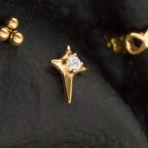 May include: A collection of gold-toned earrings. Includes a cluster of three small balls, a star-shaped stud with a central gem, and a continuous hoop design. The earrings are displayed against a dark, textured background.