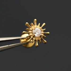 Peut inclure: Boucle d'oreille en forme de soleil de couleur or, tenue par une pince à épiler sur fond gris foncé. Le motif en forme d'éclat de soleil présente une pierre précieuse centrale et transparente. La boucle d'oreille est petite et délicate.