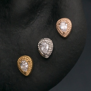 Può includere: Una collezione di piercing all'orecchio con tre borchie a forma di pera con pietre chiare centrali e bordi testurizzati color oro e argento, insieme a due cerchi color oro semplici e un piccolo perno con ciondolo color oro su un orecchio scuro.