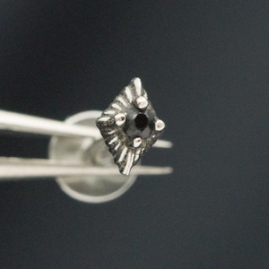 May include: A small, diamond-shaped stud earring featuring a central black stone set in a textured, silver-toned metal frame with radiating etched lines. The earring has a flat, circular backing and is held by metal tweezers for display.