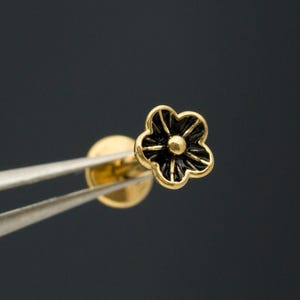 Peut inclure: Une petite boucle d'oreille à tige en or en forme de fleur à cinq pétales avec des détails en émail noir dans les zones en creux. La boucle d'oreille présente un fermoir circulaire plat et est tenue par une pince métallique sur fond sombre.
