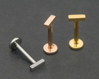 Puede incluir: Tres piezas de joyería corporal tipo labret con parte posterior plana en tonos plata, oro rosa y oro. Cada pieza presenta un poste recto con una base circular plana y una parte superior de barra rectangular horizontal para piercings.