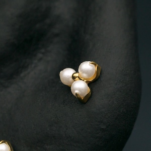 May include: Close-up of two gold earrings. One earring features a cluster of three pearls, while the other is a flower shape with pearls and a central diamond. The earrings are set against a dark background.