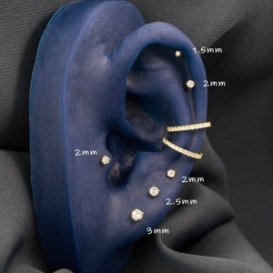 May include: A close-up of a blue ear model with multiple gold earrings with small diamonds. The earrings are labeled with their sizes in millimeters: 1.5 mm, 2 mm, 2.5 mm, and 3 mm.