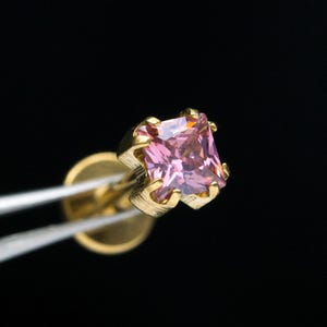 May include: A close-up of a gold-colored earring with a square, pink gemstone. The earring is held by tweezers against a black background. The gemstone is a light pink color and is set in a gold-colored metal.