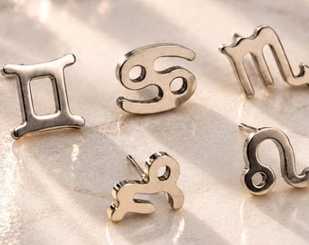 May include: Five silver-toned stud earrings shaped as zodiac symbols, including Gemini, Cancer, Scorpio, Capricorn, and Leo. Each earring features a polished metallic finish and a standard post backing, arranged together on a smooth surface.