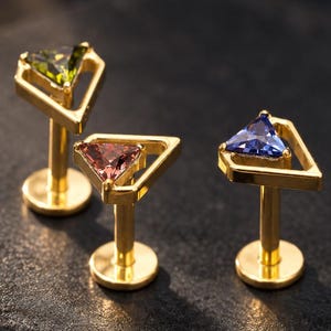 Pode incluir: Três brincos labret dourados com engastes triangulares. Cada brinco contém uma pedra preciosa de cor diferente: uma verde-oliva, uma rosa-avermelhada e uma azul. As peças de joalharia têm bases circulares planas e hastes retas.