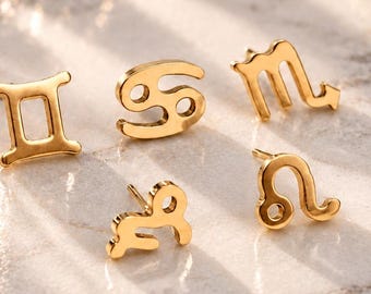 May include: Five gold-toned stud earrings shaped as zodiac symbols, including Gemini, Cancer, Scorpio, Capricorn, and Leo. Each earring features a polished metallic finish and a post back, arranged in a cluster on a smooth, light-colored surface.