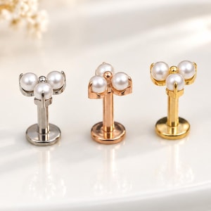 Könnte beinhalten: Drei Labret-Stecker mit flacher Rückseite, die mit einer Gruppe aus drei kleinen weißen Perlen in dreieckiger Anordnung verziert sind. Die Stecker sind in Silber-, Roségold- und Goldtönen gehalten und haben jeweils eine runde Basis.