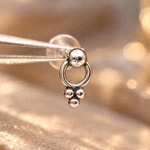 May include: A silver-toned stud earring featuring a circular ring design suspended from a small sphere, with three additional tiny spheres arranged in a triangular cluster at the bottom of the ring, held by metal tweezers.