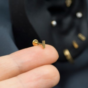 May include: A gold bar stud earring, held between two fingers.