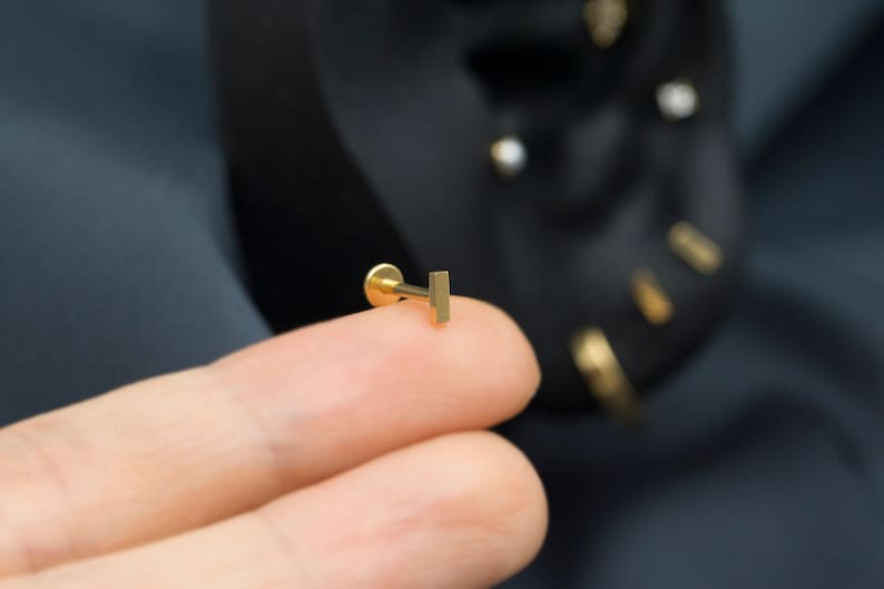 May include: A gold bar stud earring, held between two fingers.