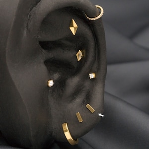 May include: A close-up of a black ear model with multiple gold earrings. The earrings include a small diamond-shaped stud, a small round stud, a small square stud, a small hoop with a row of small diamonds, and three small gold bars.
