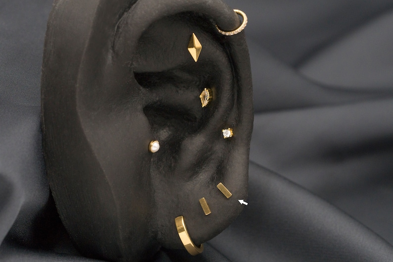 May include: A close-up of a black ear model with multiple gold earrings. The earrings include a small diamond-shaped stud, a small round stud, a small square stud, a small hoop with a row of small diamonds, and three small gold bars.
