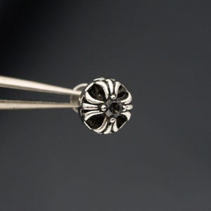 May include: A silver stud earring with a floral pattern and a black gemstone at its centre. The earring is held by silver tweezers against a dark grey backdrop. The design features curved lines radiating from the central stone.