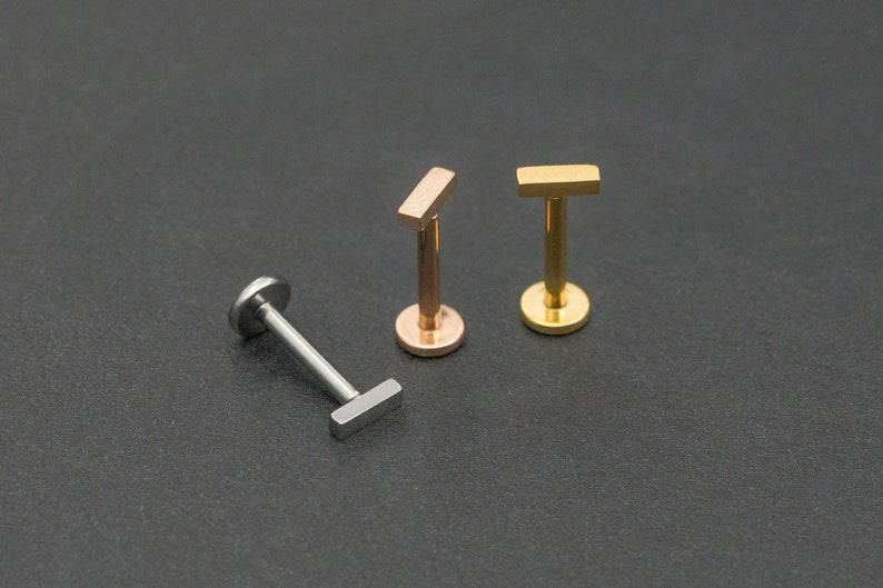 May include: Three body jewellery pieces in different colours. The jewellery is made of metal and has a small rectangular bar. The jewellery is on a black background.