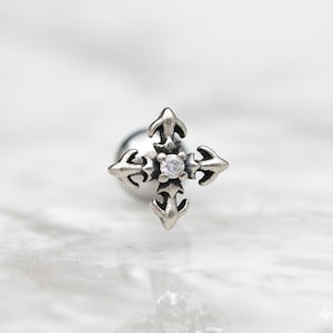 May include: A silver-toned earring featuring a cross design with four arrow-shaped points. The center of the cross holds a small, clear gemstone. The earring has a post backing.