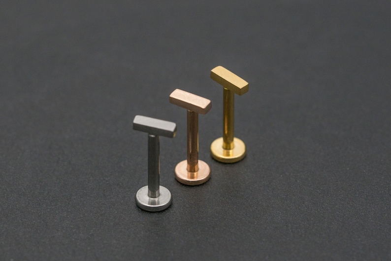 May include: Three barbell earrings with different coloured tops. The tops are silver, rose gold, and gold. The earrings are all the same size and shape.