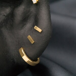 May include: A close-up of a person's ear with three gold bar earrings. The earrings are in the shape of a rectangle and are placed in the upper earlobe, helix, and conch.