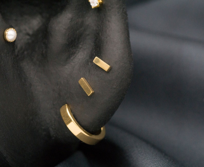 May include: A close-up of a person's ear with three gold bar earrings. The earrings are in the shape of a rectangle and are placed in the upper earlobe, helix, and conch.