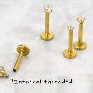 May include: Four gold-toned body jewelry pieces with clear gem accents. Each piece has a flat base and a post with a gem on the top. The text "*Internal threaded" is written on the surface below the jewelry.