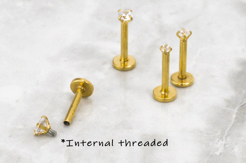 May include: Four gold-toned body jewelry pieces with clear gem accents. Each piece has a flat base and a post with a gem on the top. The text "*Internal threaded" is written on the surface below the jewelry.