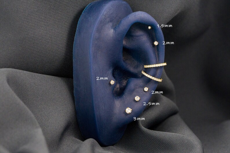 May include: A close-up of a blue ear model with multiple gold earrings with small diamonds. The earrings are labeled with their sizes in millimeters: 1.5 mm, 2 mm, 2.5 mm, and 3 mm.