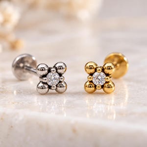 May include: Two flat back stud earrings featuring a four-petal flower design. One stud is silver-toned metal and the other is gold-toned metal. Each earring has a central clear round gemstone surrounded by four small polished metal spheres.