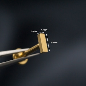 May include: Gold bar stud earring measuring 4 mm in length and 1 mm in width. The earring is being held by tweezers.