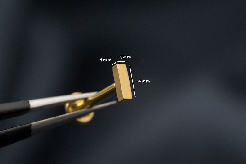 May include: Gold bar stud earring measuring 4 mm in length and 1 mm in width. The earring is being held by tweezers.