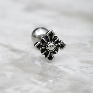 Op de afbeelding: Een zilveren oorbel met een zwart bloemenkruisontwerp. De oorbel heeft een kleine, ronde, zilveren bal aan de achterkant. Het kruisontwerp heeft een zilveren middelpunt en vier gestileerde bloemblaadjes.