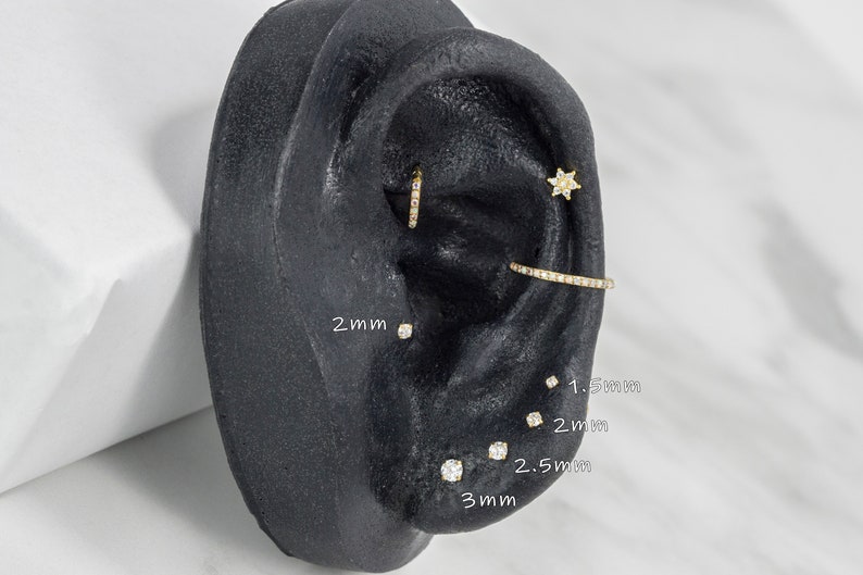 May include: A close-up of a fake ear with various sizes of diamond earrings. The earrings are 1.5mm, 2mm, 2.5mm, and 3mm in size. The ear is made of black silicone and is on a white background.