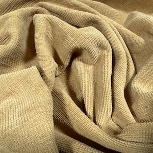 May include: Close-up of a tan corduroy fabric with a textured, ribbed surface. The fabric is draped and folded, creating shadows and highlights that emphasize its texture. The material appears soft and pliable.